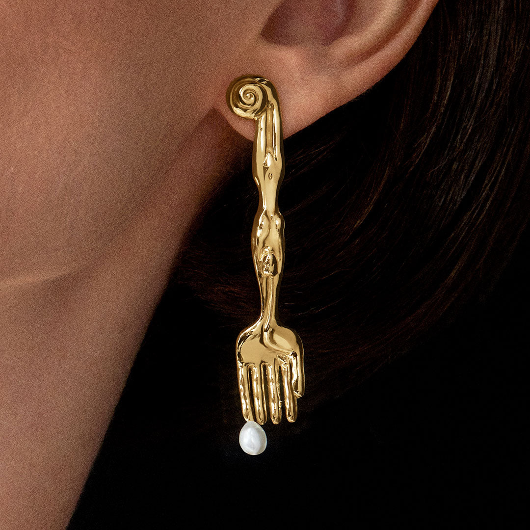 Model wearing sterling silver earring with 18k gold plating in the shape of a fork with surrealist body and natural pearl