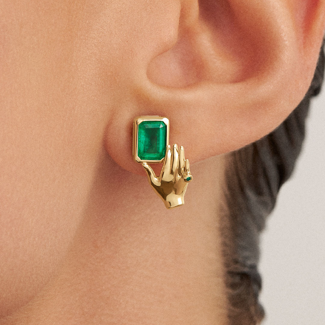 Model wearing 18k gold earring of a hand holding a rectangular emerald