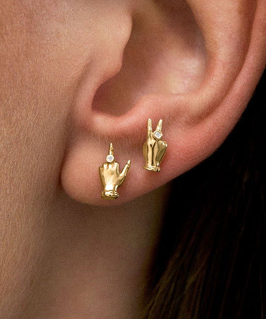 Model wearing 18k gold earrings of mini hands in peace and middle finger gestures with diamonds