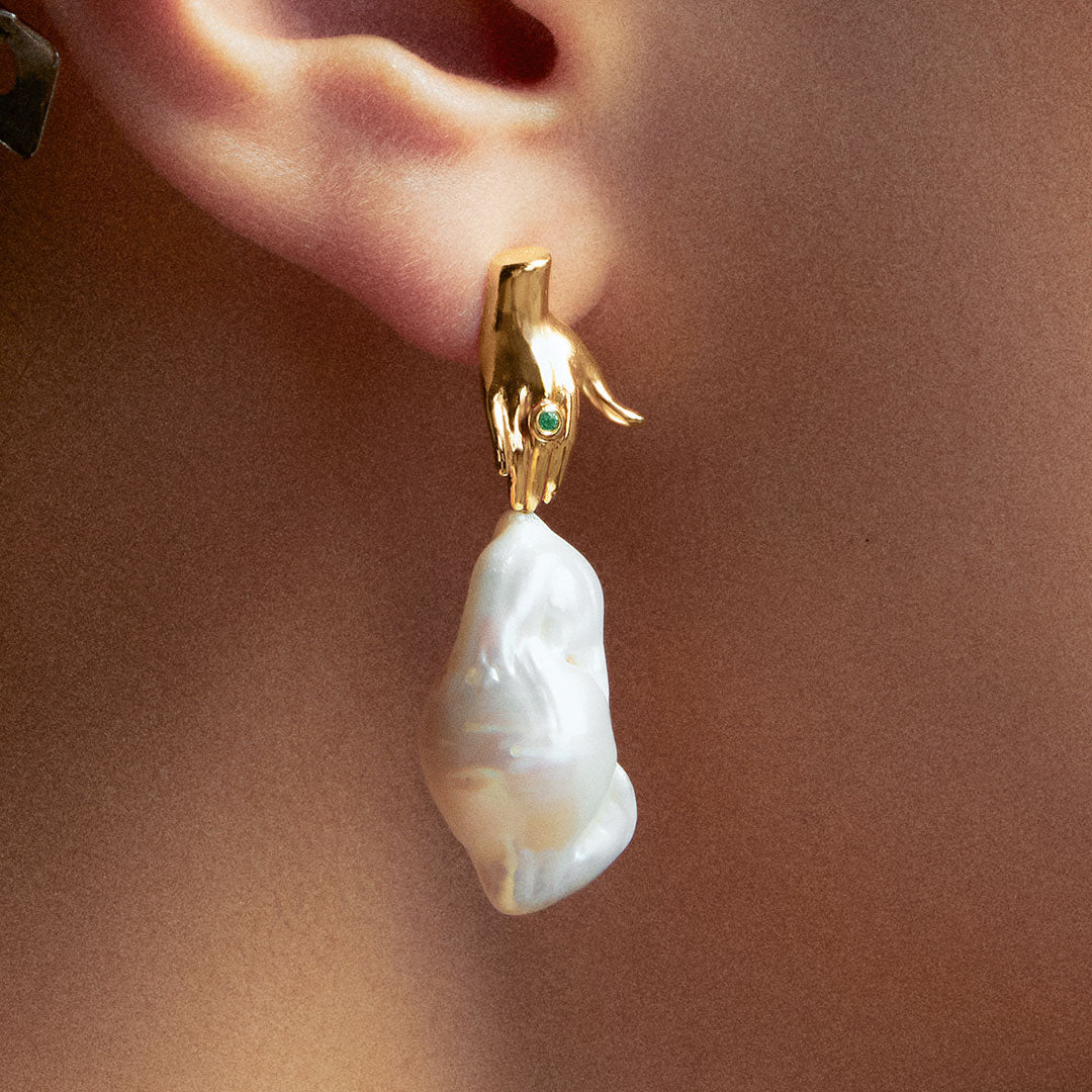 Model wearing hand-shaped earring in sterling silver with 18k gold plating, emerald, and baroque pearl