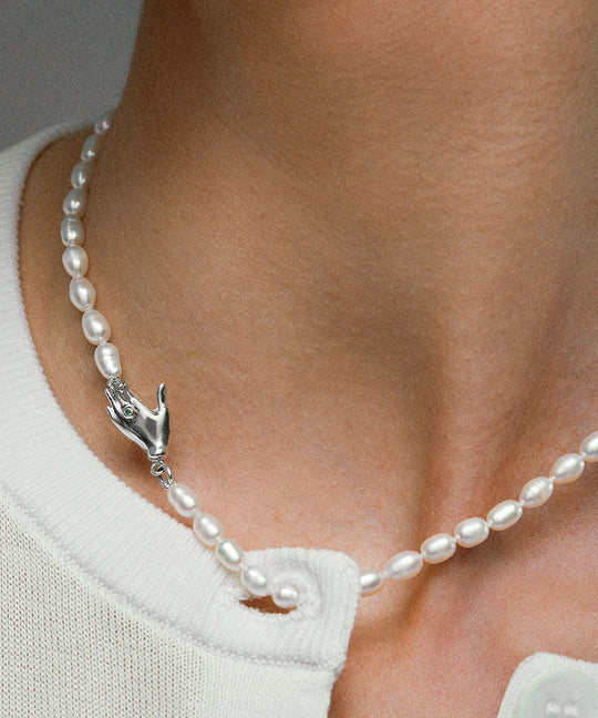 Model wearing pearl necklace with hand-shaped clasp in sterling silver adorned with emerald