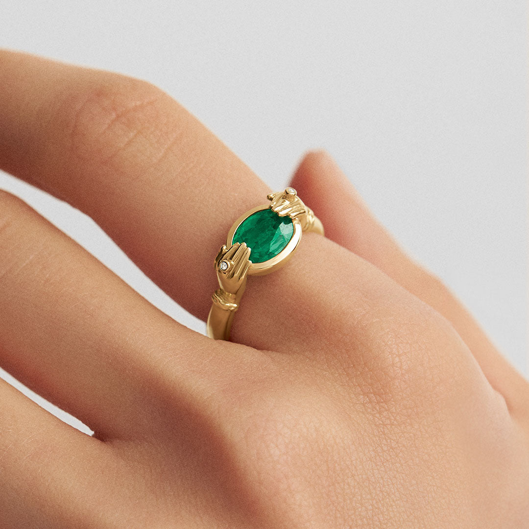 Model wearing 18k gold ring of hands adorned with diamonds holding an oval emerald
